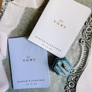 May include: A white vow book with gold text reading her VOWS and a light blue vow book with gold text reading his VOWS, both personalized with HANNAH & JONATHAN 06.10.23. A blue velvet hexagonal ring box contains two rings, accompanied by a light pink ribbon.