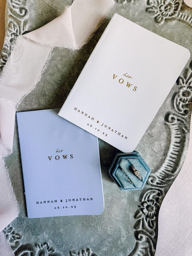 May include: A white vow book with gold text reading her VOWS and a light blue vow book with gold text reading his VOWS, both personalized with HANNAH & JONATHAN 06.10.23. A blue velvet hexagonal ring box contains two rings, accompanied by a light pink ribbon.