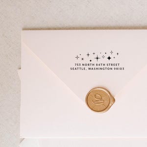 May include: A light pink envelope featuring a black ink return address that reads 753 NORTH 84TH STREET SEATTLE, WASHINGTON 98103, accented by a row of small star icons above the text and a circular gold wax seal on the bottom flap.