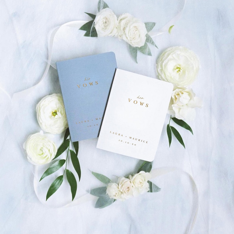 May include: A dusty blue booklet with gold text reading his VOWS and a white booklet with gold text reading her VOWS, both personalized with the names Laura and Maurice and the date 10.10.20, surrounded by white ranunculus flowers and green leaves.