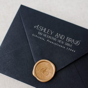 May include: A matte black envelope featuring white printed text that reads ASHLEY AND BRAD, 199 MOUNTAIN VIEW DRIVE, Gibsonia, Pennsylvania 15044. The envelope is sealed with a circular metallic gold wax seal embossed with a cursive letter B.