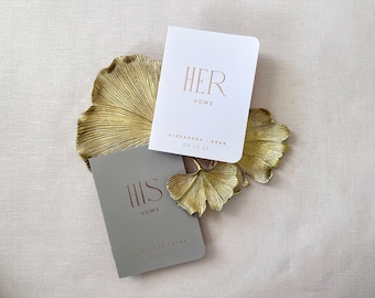 May include: Two vow books, one white and one gray, rest on a gold leaf-shaped tray. The white book reads "HER VOWS" with the names and date below. The gray book reads "HIS VOWS" with names and date below. Wedding theme.