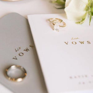 May include: A gray booklet with the text his VOWS and a white booklet with the text her VOWS in gold lettering. A gold engagement ring with a rectangular stone rests on the white booklet, and a plain gold band rests on the gray booklet.