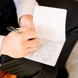 May include: A small white notebook with lined pages featuring handwritten black ink text. The notebook is held open while a black ballpoint pen is used to write on the paper. The visible text begins with the name Olivia and continues with sentences.