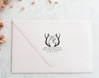 May include: A light pink envelope featuring a black ink stamp design of two deer antlers framing the initials J & K. Below the antlers, the text reads 1928 Presidential Drive, Allison Park, Pennsylvania, 1-5-1-6-1 in a classic serif font.
