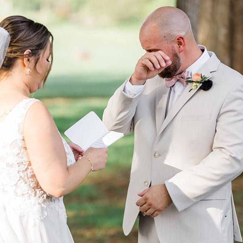 May include: A white sleeveless wedding dress with floral lace details and a beige suit jacket with a patterned bow tie, a peach-colored boutonniere, and a white pocket square. The suit jacket features two buttons and a notched lapel design.