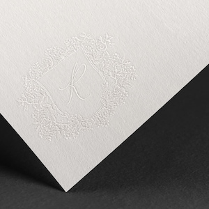 May include: A white card with a white embossed design of a floral wreath with the letter "R" in the center.
