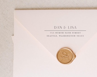 May include: A pale pink envelope featuring black printed text that reads DAN & LISA, 753 NORTH 84TH STREET, SEATTLE, WASHINGTON 98103. The envelope is sealed with a circular gold wax seal embossed with a stylized letter G in the center.