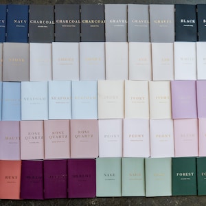 May include: A collection of rectangular notebooks arranged in a grid, featuring various solid colors including navy, charcoal, black, white, mauve, rose quartz, rust, merlot, sage, and forest green, each stamped with metallic foil text.