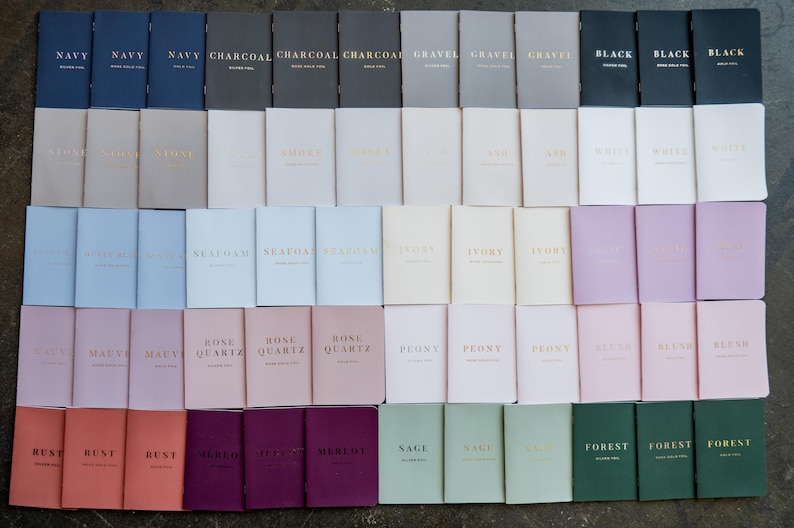 May include: A collection of rectangular notebooks arranged in a grid, featuring various solid colors including navy, charcoal, black, white, mauve, rose quartz, rust, merlot, sage, and forest green, each stamped with metallic foil text.