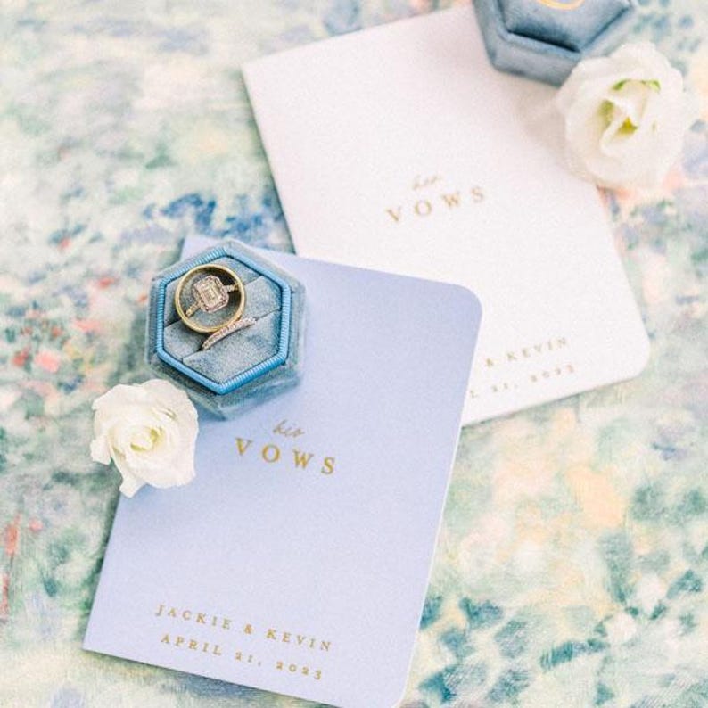 May include: A light blue velvet ring box containing a gold engagement ring with a rectangular stone, placed on a light blue vow book with gold text reading his VOWS, JACKIE & KEVIN, APRIL 21, 2023, accompanied by a white vow book and white flowers.