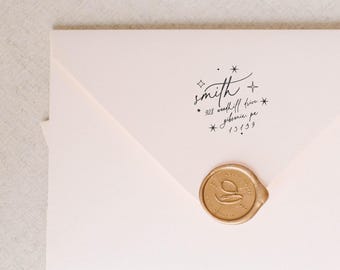 May include: A pale pink envelope featuring a black ink return address stamp that reads smith, 928 woodhill drive, gibsonia, pa, 15139, accented with small star icons. A circular gold wax seal with an embossed letter G is centered on the envelope flap.