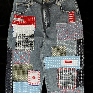 May include: Blue denim jeans with a patchwork design featuring various colors and textures, including red, white, and blue. The jeans have a drawstring waist and are decorated with intricate stitching.