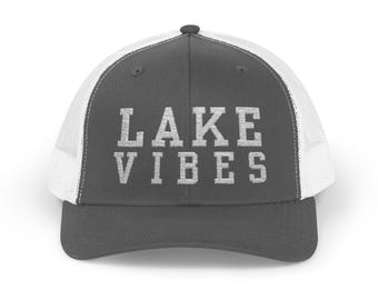 May include: A gray trucker hat with a white mesh back and a curved brim. The hat features the text "LAKE VIBES" in bold, white letters across the front. The hat is designed for outdoor activities.