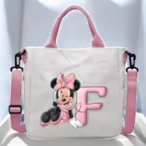 Puede incluir: Una bolsa de lona blanca con asas rosas y una correa de hombro ajustable rosa. La bolsa presenta una imagen de dibujos animados de Minnie Mouse con un lazo rosa y la letra "F" en rosa.