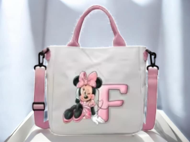 Puede incluir: Una bolsa de lona blanca con asas rosas y una correa de hombro ajustable rosa. La bolsa presenta una imagen de dibujos animados de Minnie Mouse con un lazo rosa y la letra "F" en rosa.