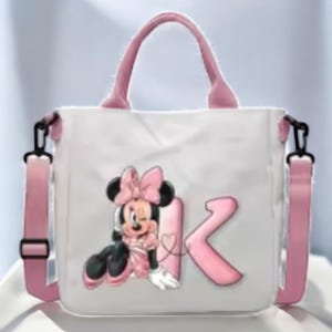 Puede incluir: Una bolsa de lona blanca con asas rosas y una correa de hombro rosa. La bolsa presenta una imagen de dibujos animados de Minnie Mouse con un lazo rosa y la letra "K" en rosa.
