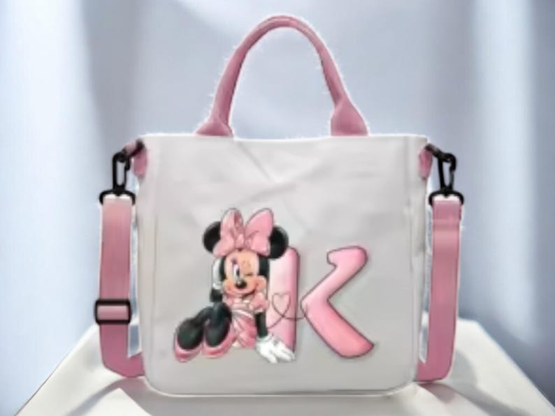 Puede incluir: Una bolsa de lona blanca con asas rosas y una correa de hombro rosa. La bolsa presenta una imagen de dibujos animados de Minnie Mouse con un lazo rosa y la letra "K" en rosa.