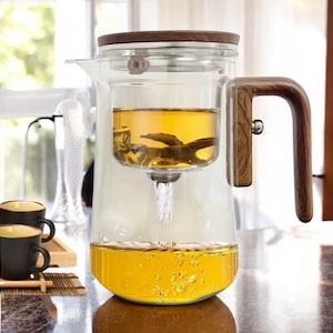 Puede incluir: Una tetera de vidrio con asa y tapa de madera. La tetera tiene un infusor incorporado y está llena de té amarillo.