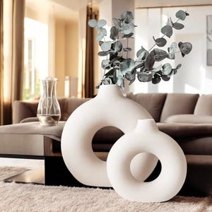 Puede incluir: Dos jarrones de cerámica blanca con una abertura en forma de anillo. Los jarrones están sobre una mesa negra con una alfombra marrón debajo. Un ramo de ramas de eucalipto está en el jarrón más grande.