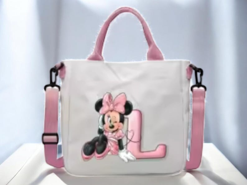 Puede incluir: Una bolsa de lona blanca con correas rosas y un personaje de Minnie Mouse rosa y negro con la letra "L" en la parte delantera.