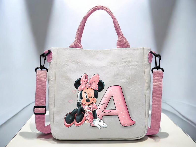 Puede incluir: Bolso de lona rosa con un dise&ntilde;o de personaje de Minnie Mouse. Minnie lleva un vestido y un lazo rosa y sostiene una letra A rosa. El bolso tiene una correa de hombro rosa.