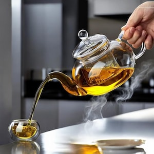 May include: A clear glass teapot with a glass lid is pouring tea into a small glass cup. The tea is a golden colour, and steam rises from the cup and teapot. The teapot is held by a hand.