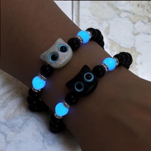 Pode incluir: Duas pulseiras que brilham no escuro com contas pretas e pingentes de gato. Um pingente é branco com olhos azuis e o outro é preto com olhos azuis.