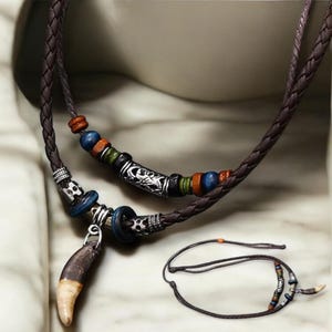 Puede incluir: Un collar de cuero marrón con un colgante de diente y cuentas de colores. El collar está trenzado y tiene un dije de plata.