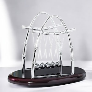 Puede incluir: Un péndulo de Newton de metal plateado con cinco bolas de metal suspendidas de un marco cromado. Las bolas están suspendidas de una base de madera con un acabado marrón oscuro.