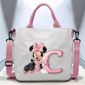 Puede incluir: Una bolsa de lona blanca con asas rosas y un dise&ntilde;o de Minnie Mouse rosa y negro. El dise&ntilde;o presenta a Minnie Mouse con un lazo rosa, gui&ntilde;ando un ojo, y sentada junto a una gran letra rosa "C".