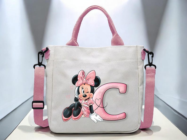 Puede incluir: Una bolsa de lona blanca con asas rosas y un dise&ntilde;o de Minnie Mouse rosa y negro. El dise&ntilde;o presenta a Minnie Mouse con un lazo rosa, gui&ntilde;ando un ojo, y sentada junto a una gran letra rosa "C".