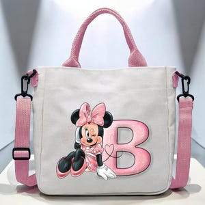 Puede incluir: Una bolsa de lona blanca con correas rosas y un personaje de Minnie Mouse rosa y negro con la letra "B" en purpurina rosa.