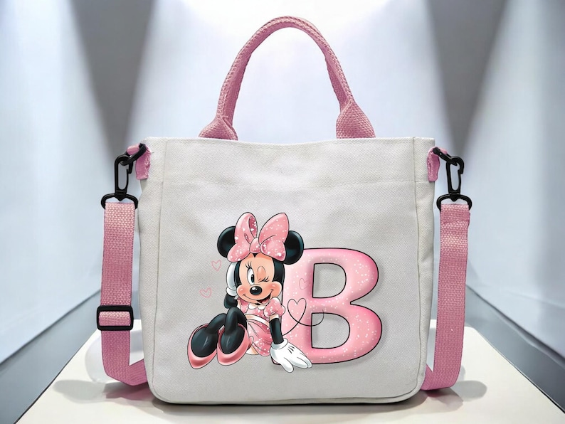 Puede incluir: Una bolsa de lona blanca con correas rosas y un personaje de Minnie Mouse rosa y negro con la letra "B" en purpurina rosa.