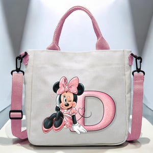 Puede incluir: Bolso tote de lona rosa con un personaje de Minnie Mouse rosa y blanco y la letra D. El bolso tiene una correa de hombro rosa y asas negras.