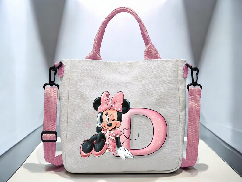 Puede incluir: Bolso tote de lona rosa con un personaje de Minnie Mouse rosa y blanco y la letra D. El bolso tiene una correa de hombro rosa y asas negras.