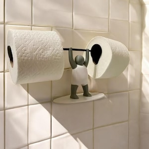 Könnte beinhalten: Ein weißer Toilettenpapierhalter mit einer grauen Figur eines Menschen, der eine Langhantel hebt. Der Halter ist an einer weißen gekachelten Wand montiert.
