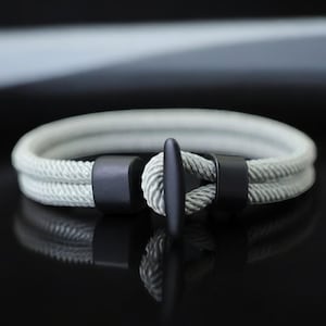 May include: A gray rope bracelet with a black anchor clasp. The bracelet is made of two strands of rope and has a simple design.
