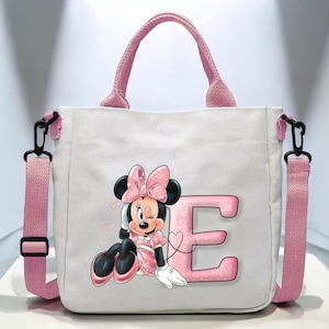Puede incluir: Una bolsa de lona rosa con una correa de hombro negra. La bolsa presenta una letra "E" rosa y una imagen de dibujos animados de Minnie Mouse con un lazo rosa y un vestido rosa.
