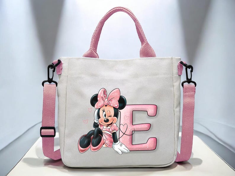 Puede incluir: Una bolsa de lona rosa con una correa de hombro negra. La bolsa presenta una letra "E" rosa y una imagen de dibujos animados de Minnie Mouse con un lazo rosa y un vestido rosa.