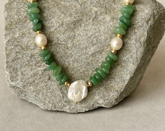 May include: A necklace with green jade chips and large, off-white pearls. The necklace has gold-coloured accents and a large, irregularly shaped pearl pendant. The jewellery is displayed on a grey stone surface.