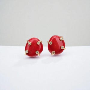 Könnte beinhalten: Paar rote, ovale Ohrstecker. Jeder Ohrring hat eine polierte, glänzende Oberfläche und ist mit kleinen, goldfarbenen Zinken verziert, die klare, funkelnde Steine halten. Die Ohrringe sind vor einem weißen Hintergrund platziert.
