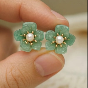 Può includere: Un paio di orecchini a forma di fiore. Ogni orecchino presenta un fiore di giada verde chiaro con cinque petali, una perla centrale e dettagli color oro. Gli orecchini sono tenuti nella mano di una persona.