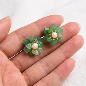 May include: A pair of flower-shaped earrings. Each earring features a cluster of green stones forming petals around a central pearl. The earrings have gold-coloured accents and are displayed on a light background.
