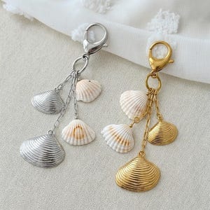 Op de afbeelding: Twee sleutelhangers met schelpenbedels. De ene sleutelhanger is zilverkleurig met zilveren schelpenbedels en de andere is goudkleurig met gouden schelpenbedels. De schelpen zijn wit en bruin.