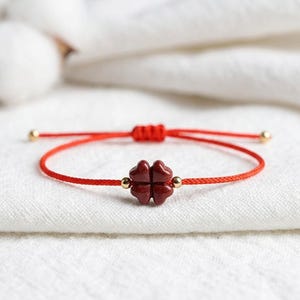 May include: A red string bracelet with a dark red four-leaf clover charm. The bracelet has gold-coloured beads and a sliding knot closure. The bracelet is displayed on a white fabric background.