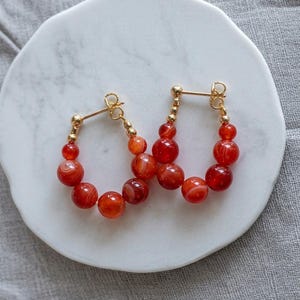 May include: A pair of hoop earrings featuring round, translucent orange and red agate beads accented with small gold-toned metal spheres. The earrings have a gold-toned post back closure and are arranged in a circular shape on a white surface.