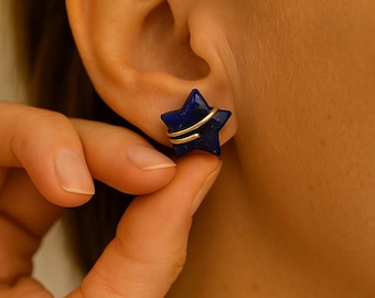 Könnte beinhalten: Ein dunkelblauer, sternförmiger Ohrring mit einer goldfarbenen Drahtwicklung. Der Ohrring wird an einem Ohr gehalten, um das Design und die Farbe des Schmucks zu zeigen. Der Ohrring besteht aus einem dunkelblauen Stein.