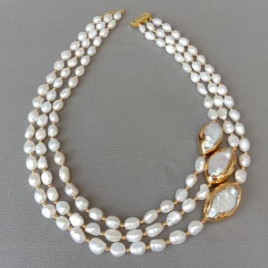 Peut inclure: Un collier de perles à trois rangs avec un fermoir et des accents dorés. Le collier présente des perles blanches de forme ovale et trois perles plus grandes, de forme irrégulière, avec des bordures dorées.