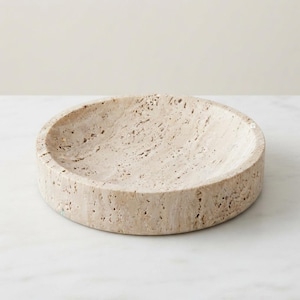 May include: A circular, shallow bowl crafted from beige travertine stone. The surface features the natural, porous, and pitted texture characteristic of travertine, with a consistent light tan colour throughout the entire round object.